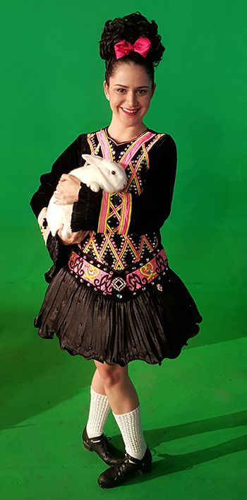 Victoria Molina wearing her Irish step dancing costume: a wig with cascading jet black curls, an elaborately bedazzled dress, and high white socks.