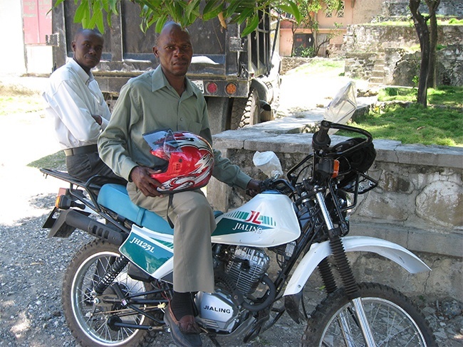 Two of the supervisors of schools in Port-de-Paix, Haiti, Miami's sister diocese sit atop the Chinese-made motorbike that will help them visit schools and train teachers in the remote mountainside schools.