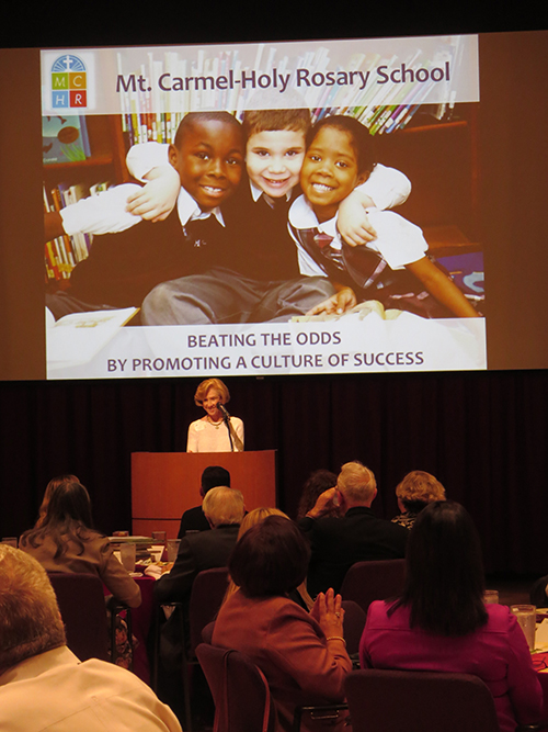 Wini Amaturo speaks at the luncheon honoring the Amaturo Foundation's decade of support for Catholic schools in South Florida. Behind her is a slide showing the Harlem school whose principal spoke at the luncheon.