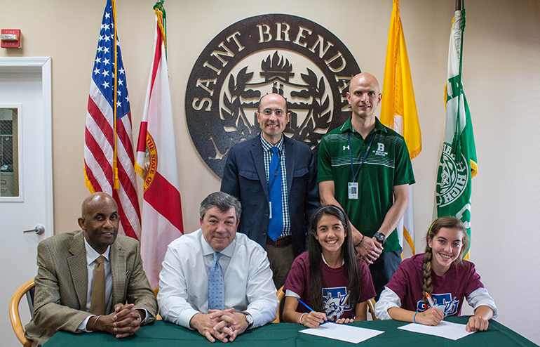 Adriana Prieto, seated second from right, and Nikki Dennis, far right, sign their Letter of Intent to play soccer at St. Thomas University. Seated next to them are soccer coaches Mike Werner and Bert Jordan. Standing behind them, from left, are school principal Jose Rodelgo-Bueno and Julio Arnaiz, athletic director.