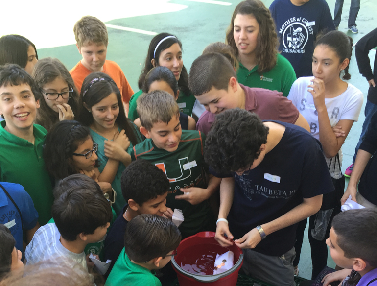 With a mix of middle, high and college student minds in action at STEM Day at St. Brendan High, students tested out the "boats" they had to design to carry as many pennies as possible. FIU student Pedro da Costa (center in navy blue) placed the test paper boat in water and students counted how many pennies it could hold.