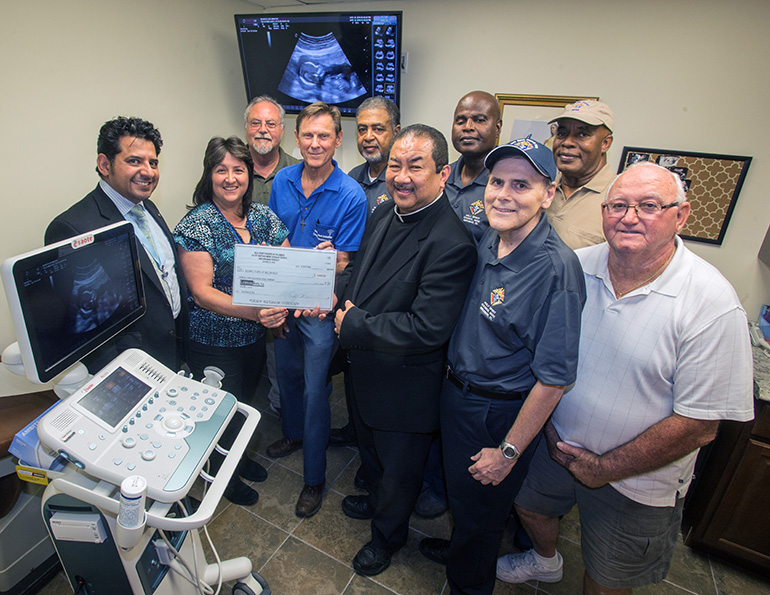 Posing for a photo with an enlarged version of a ,000 check presented to the South Broward Pregnancy Help Center, from left, are: Jorge Ibacache; Sandi Le Bel, the center's program coordinator; Clifton Day; Charlie Fitch; Anthony Dyett; Father Andrew Chan-A-Sue; pastor of St. Bartholomew Church in Miramar; Michael Thomas, Grand Knight of the Knights of Columbus Holy Spirit Council 6032; John Venezia; Tracy King; and Orville Wade The men are all members of the Knights' Council 6032. A picture of an unborn baby appears above them on the TV screen, and the ultrasound machine is in the foreground.