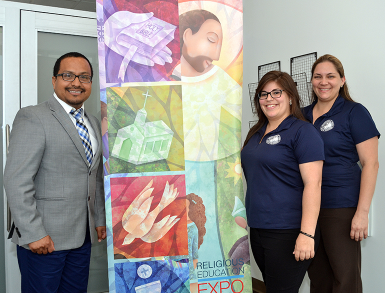 Peter J. Ductram, director of catechesis for the archdiocese, shows the banner of the recent Religious Education Expo. On the right are his staffers: administrative assistant Yomaira Diaz (next to the banner) and Teresita Prieto, coordinator of formation and certification.