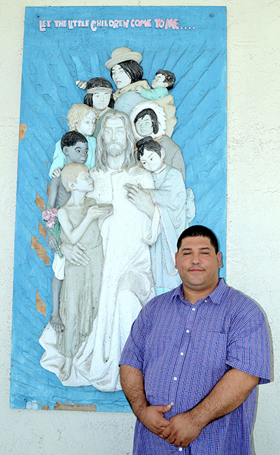 Pablo Cuadra, director de formación religiosa en el Schott Center, frente al bajorrelieve de Jesús con los niños. La obra está situada a la entrada de St. Jude Chapel, en los terrenos del Schott Center.