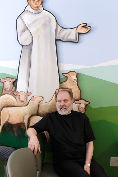 At Camillus House, Hospitaller Brother Mateo Fenza poses in front of the mural of the "lost sheep" in the chapel, one of the new Camillus buildings. The former soup kitchen now includes a behavioral health treatment and residential facility, medical and service space, dining hall and cafe, parking garage, commercial kitchen, offices, educational spaces, and an auditorium.