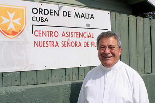 Mons. Tom&aacute;s Mar&iacute;n, capell&aacute;n de la Asociaci&oacute;n Cubana de la Orden de Malta, se retrata en frente del letrero que anuncia uno de sus proyectos en Miami, la Casa de Malta en la parroquia San Juan Bosco de la Peque&ntilde;a Habana.