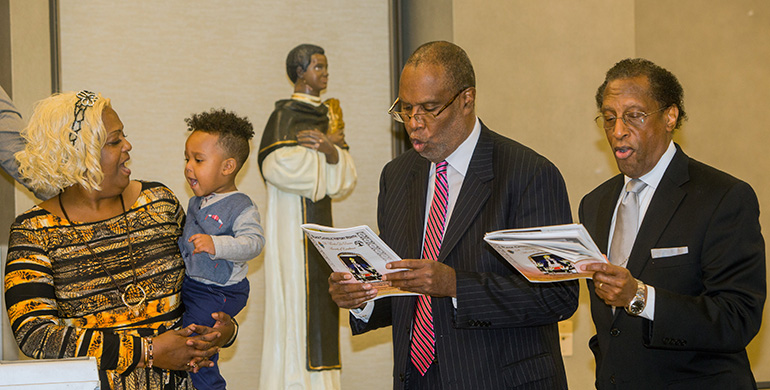 Joziah Alago, 18 months old, held by Peggy McIntyre, joins Marvin Dawkins, University of Miami sociology professor, and Don Edwards, associate superintendent of Archdiocese of Miami schools, as they sing, "Lift Every Voice And Sing," at the St. Martin de Porres awards luncheon.