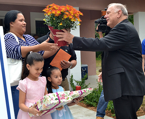 El Arzobispo Wenski le entrega flores a Isela Calderón como regalo de inauguración de la Casa Papa Francisco II, mientras sus hijas Elizabeth, de rosado, y Jennifer miran con entusiasmo las cajas de regalo que les dieron.