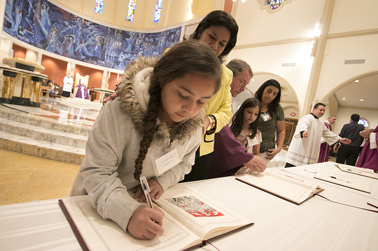 La catecúmena Mya Matamoros, de 10 años, de la parroquia St. Martha,  en Miami Shores, firma en el Libro de los Elegidos, mientras su madrina, Dannys Cabrera observa durante la primera de dos ceremonias del Rito de la Elección, el 5 de marzo, en la Catedral St. Mary.