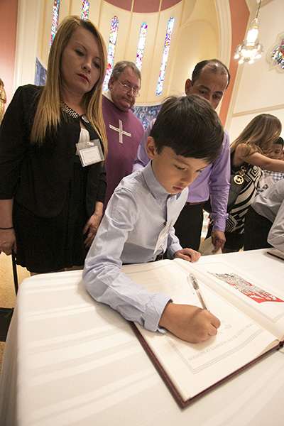 Catechumen Julian David Leon Mora, 9, signs his name in the Book of the Elect as her godparents, Nohora Astrid Mora and Nelson Gustavo Crispin of St. Martha Parish in Miami Shores, look on, during the first of two Rite of Election ceremonies March 5 at St. Mary Cathedral.