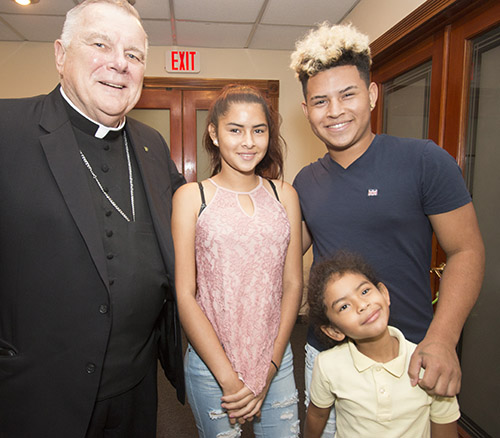 El Arzobispo Thomas Wenski posa con los hermanos Romel Cardoza, de 16 años; Lizamary Cardoza, de 14 años; y Vanessa Cardoza, de 5 años, durante una visita a las oficinas de Catholic Legal Services de Miami, el 27 de septiembre, para lanzar la campaña mundial del Papa Francisco “Compartiendo el viaje” en favor de los migrantes y refugiados. Los adolescentes están tratando de ajustar su estatus migratorio después de llegar a Estados Unidos como menores no acompañados, hace dos años para reunirse con su madre.