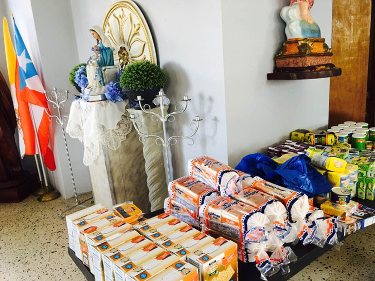 Photo of food and other provisions purchased by members of San Francisco Javier Parish in Barrio Navarro, Gurabo, Puerto Rico, for sharing with the community. At rear, the flags of the Vatican and Puerto Rico and the image of the island's patroness, Our Lady of Divine Providence.