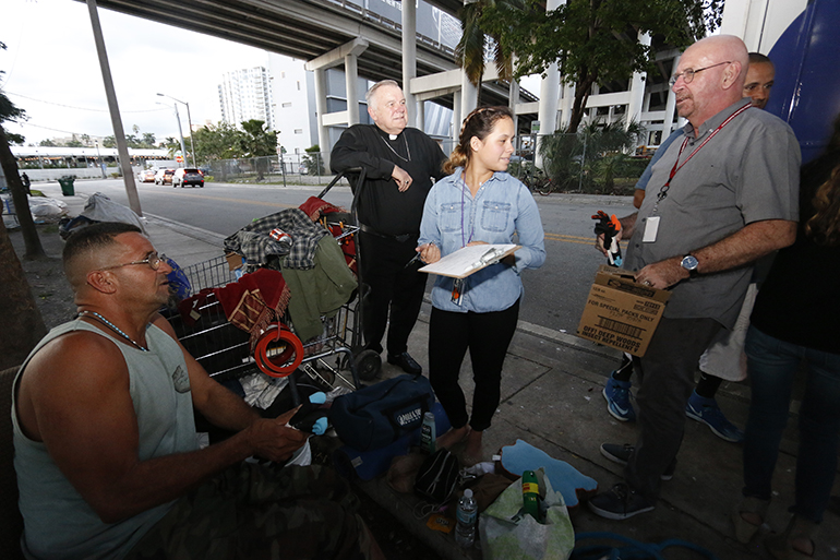 Roberto Zuñet, seated, speaks with Hospitaller Brother Richard Moore as Lazarus Project intern Gabriela Puig and Archbishop Thomas Wenski stand by.