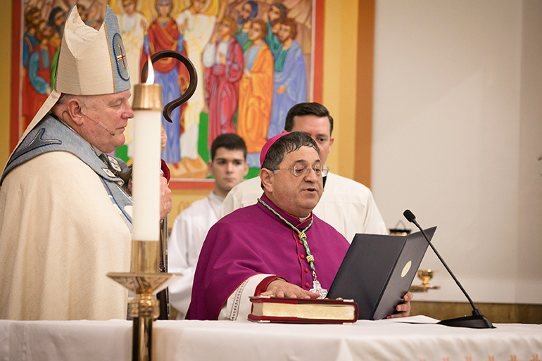 Bishop-elect Enrique Delgado takes an oath of fidelity and signs documents at St. Katharine Drexel Parish in Weston during solemn vespers on the eve of his ordination as the newest auxiliary bishop of the Archdiocese of Miami. At left is Archbishop Thomas Wenski, and at right is his master of ceremonies, Father Richard Vigoa.