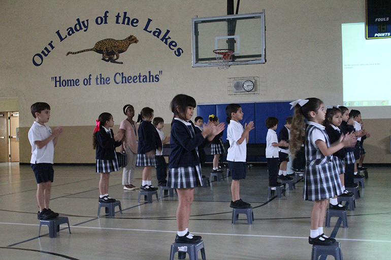 Our Lady of the Lakes School kindergarten students clap along while balancing on step stools during their NeuroNet Learning morning exercises.