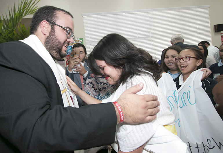 At the cathedral hall after the ceremony, Father Matthew Gomez gives one of his first blessings to Rebecca Garcia, current coordinator of Encuentros Juveniles, as fifth-graders from St. Bonaventure School in Davie, who " adopted"  him as a seminarian, cheer nearby.