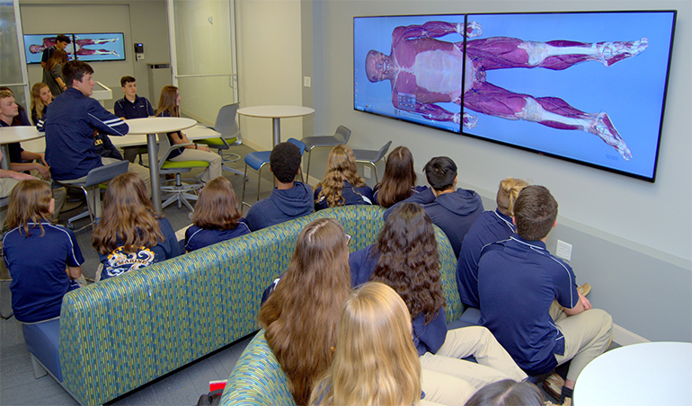 Students in the Innovation Center at St. Thomas Aquinas High School watch a full-size monitor image of a virtual cadaver, being examined by students in left background.