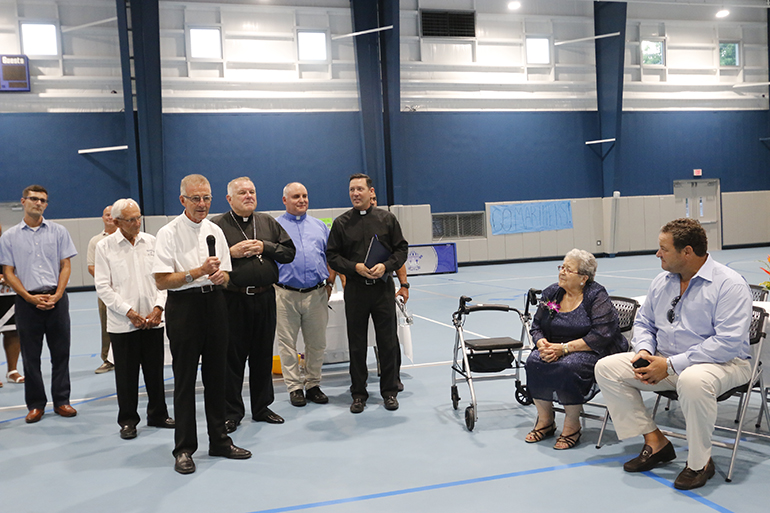 Father John Baker, rector of the Basilica of St. Mary Star of the Sea, speaks at the dedication and blessing of The Basilica School's new gym and multipurpose building, known as the Rose Renna Activity Center. Seated at right, next to her son, is Renna, whose daughter donated the $ 1 million that helped make the building happen, and named it in her mother's honor.