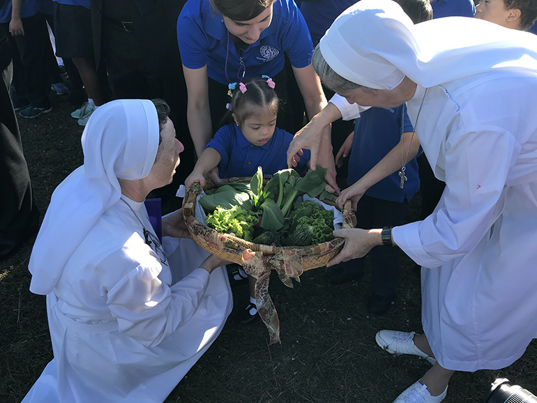 La Hna. Lidia Valli (izquierda) y la Hna. Filomena Mastrangelo, junto con el personal del Marian Center, ayudan a la estudiante, Ariana Fortuna a sostener la canasta de verduras cultivadas en la granja urbana que se le presentó al Arzobispo Thomas Wenski.