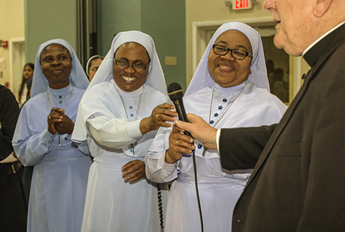 La Hna. Marygracious Onwukwe le entrega el micrófono al Arzobispo Thomas Wenski después de que ella y las hermanas Therese Martin y Mary Agatha, de las Hijas de María, Madre de la Misericordia, cantaran una bendición de los alimentos en nigeriano, en la recepción que siguió a la Misa por el Día Mundial de la Vida Consagrada.