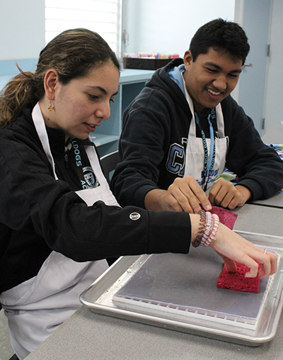 Con pantallas protectoras y absorbente, los estudiantes Briana Cruz y Nicholas Pardo eliminan el exceso de agua, la parte más larga del proceso de fabricación de papel.