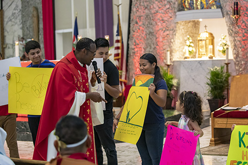 Father Robes Charles, pastor at St. Clement Church in Wilton Manors, addresses his congregation at the end of the Palm Sunday Mass. The children behind him held posters indicating their support of the Virtus program as part of the parish's Safe Environment Awareness and Prevention Family Day. Father Charles reads out-loud the message on the poster held by Jessica Alvarez, a member of St. Clement's Youth Group.