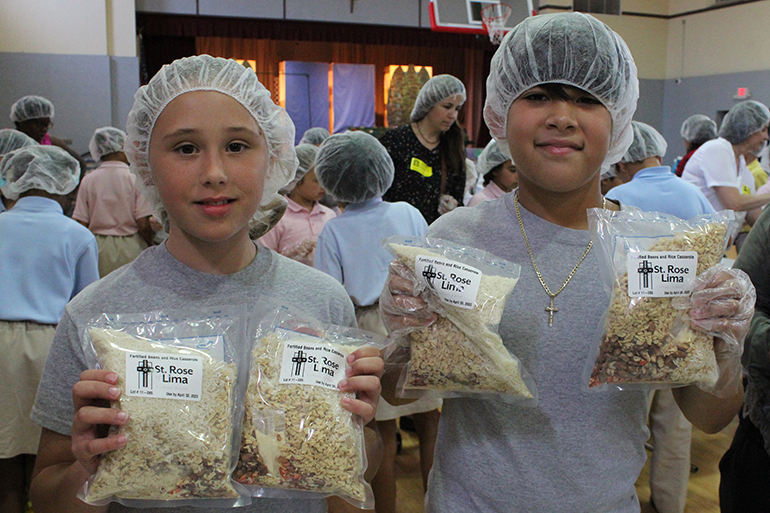 Los estudiantes de cuarto grado de la escuela St. Rose of Lima, Chloe Humbles y Evan Paul, sostienen comidas empaquetadas en el evento de empaque de comidas, Meals of Hope, en el gimnasio escolar, el 30 de abril.