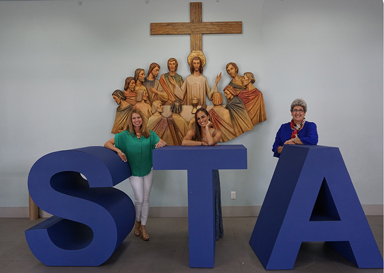 Women of STA: Principal Lisa Figueredo, alumnus Monica Puig, and teacher Nuria Baldor pose for a photo with St. Thomas the Apostle School initials. Puig, who is now an Olympic medalist and tennis pro, visited the school during the summer to highlight her early education and faith in a video for The Players' Tribune and Mass Mutual.