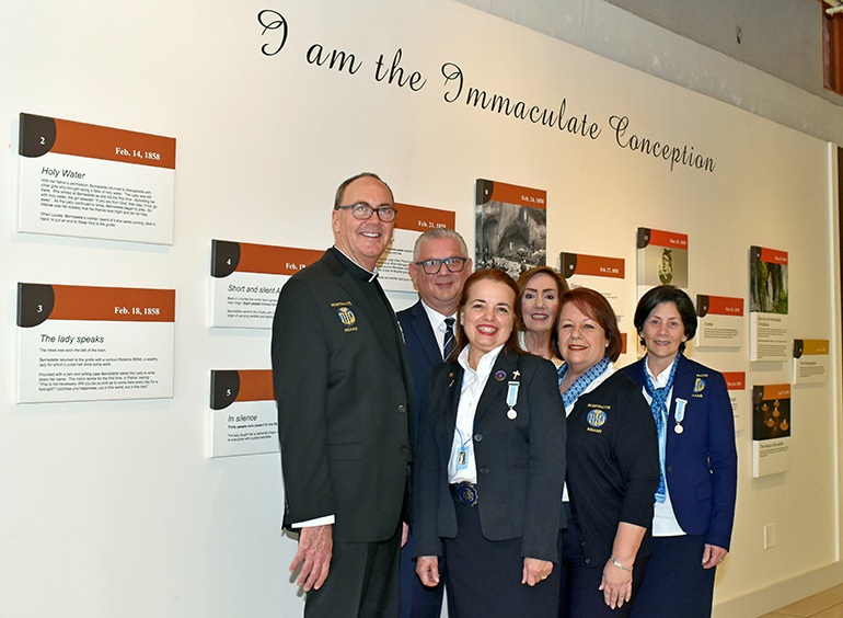 Members of Hospitalité de Miami gather at the timeline wall at Our Lady of Lourdes Church in Miami. Standing with Msgr. Kenneth Schwanger, the pastor, are, from left: Filippo Baglio, Mercy Baglio, Debra Bartkowski, Carmen Van Scoy and Fanny Garcia.