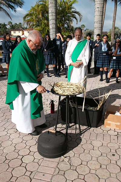 Father Juan Hernandez, pastor of Mother of Our Redeemer in Miami, lights the palms as Deacon Orlando Rojo and school children look on.