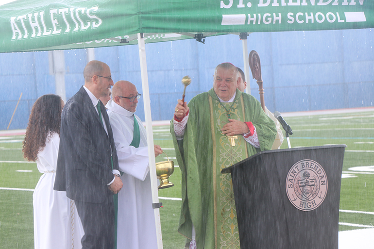 In the midst of a downpour, Archbishop Thomas Wenski blesses St. Brendan High School's new athletic fields, Feb. 19, 2020. At left are Jose Rodelgo-Bueno, the high school's principal, and Deacon Eduardo Blanco, theology teacher.