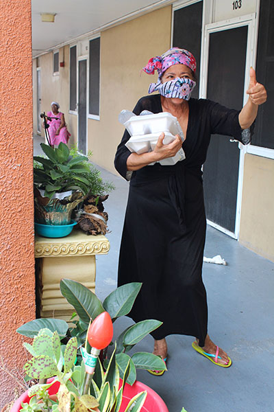 Showing off her home garden and hot meals, a resident of an apartment building in Little Haiti, wearing a mask to protect herself against COVID-19, gives a thumbs-up for the generosity of the volunteers from Notre Dame d'Haiti Mission who cooked and delivered the meals April 11, 2020.