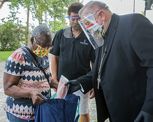 Archbishop Thomas Wenski places fish into a the bag of a food recipient as Catholic Charities CEO Peter Routsis-Arroyo looks on. The archbishop helped Father Reginald Jean-Mary, Notre Dame d'Haiti's pastor, hand out food at the church May 23, 2020.