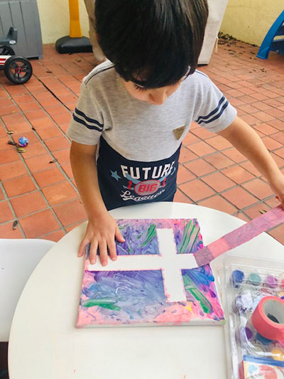 Después de pintar en un lienzo con diferentes colores, un estudiante de la Escuela St. Hugh retira la cinta en forma de cruz. Aunque las clases se realizaron en línea, después del 17 de marzo de 2020, las tareas siguieron siendo igual de prácticas y creativas.