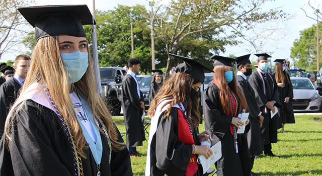 La graduada de la Secundaria Archbishop Coleman Carroll, Verónica Camacho y sus compañeros de clase se ponen de pie al inicio de la ceremonia de graduación, celebrada al aire libre en la escuela, el 30 de mayo de 2020. Debido a COVID-19, se pidió a los padres, familiares y amigos que presenciaran la ceremonia desde sus vehículos. La escuela también transmitió en vivo la ceremonia a través de Facebook.