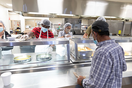 Para protegerse a sí mismos y a los demás durante la pandemia de COVID-19, los huéspedes residentes que acuden al comedor de Camillus House, en el centro de Miami, se distancian socialmente, llevan máscaras faciales y ayudan con los deberes de limpieza del refugio para indigentes. Camillus House también ha invertido mucho en lavaderos de manos y dispositivos de desinfección de alta tecnología, desplegados estratégicamente en diferentes lugares del refugio.