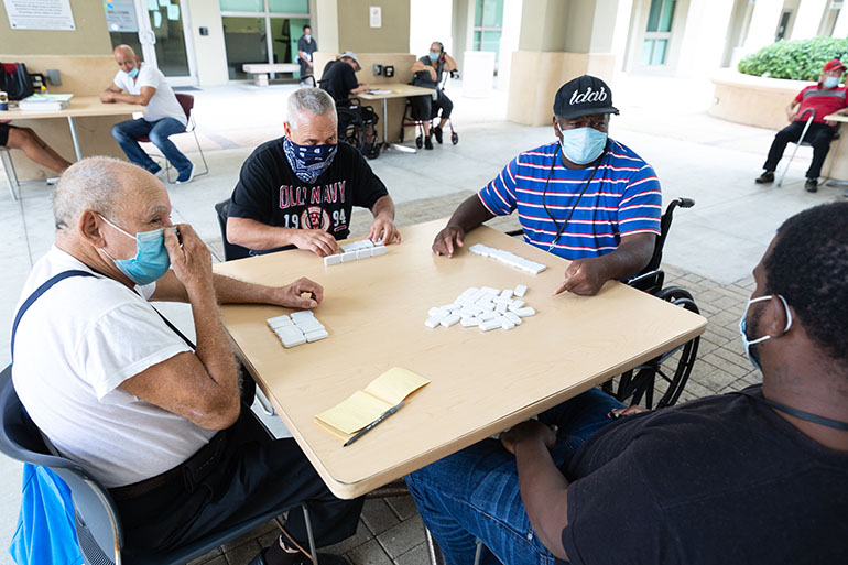 En Camillus House, en el centro de Miami, los huéspedes se divierten jugando dominó en el patio, durante la pandemia COVID-19. Para protegerse a sí mismos y a los demás, el personal y los huéspedes residentes del refugio para personas sin hogar se distancian socialmente, usan máscaras faciales y refuerzan los protocolos de saneamiento y desinfección. También están encontrando otras formas de mantener el aburrimiento a raya.