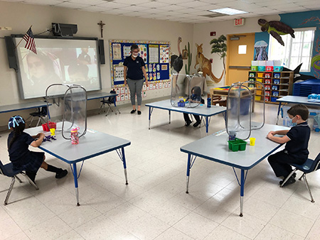 Photo of a socially distanced PreK4 classroom at Mother of Our Redeemer School, Miami, on the first day of school, Aug. 19, 2020. The in-person students are surrounded by sneeze guards at their desks, the teacher, Patricia Medina, is wearing a mask/shield, and other students are learning from home.