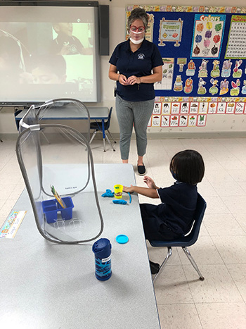 Photo of a socially distanced PreK4 classroom at Mother of Our Redeemer School, Miami, on the first day of school, Aug. 19, 2020. The in-person students are surrounded by sneeze guards at their desks, teacher Patricia Medina is wearing a mask/shield and other students are learning from home.