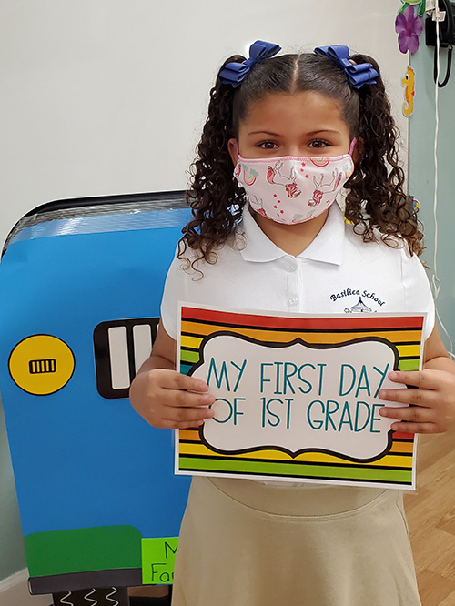 Mila Cabrera, dressed in uniform and with her hair decorated in ribbons, holds up a sign and poses for a photo on her first day back to classes at The Basilica School of St. Mary Star of the Sea in Key West.