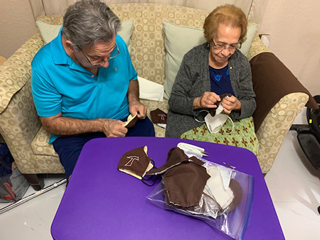 Ayudan a virar las máscaras después de ser cosidas el integrante de la fraternidad francisana seglar Espejo de Cristo, Reny Aguilera, OFS, e Hilda Nieves.