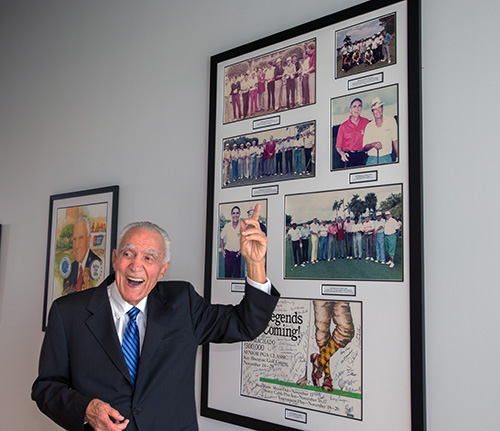 Gus Machado gestures to photos hanging on a wall in the new building from the Gus Machado Senior PGA classics he sponsored in the late 1980s. He has now given his name to the School of Business complex at St. Thomas University, dedicated Oct. 16, 2020.