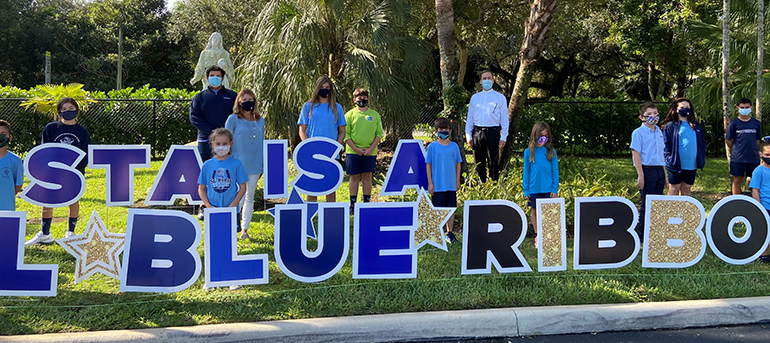 Students, principal Lisa Figueredo (middle left) and pastor, Father Alejandro Rodriguez Artola (rear right)pose amid the Blue Ribbon sign on the lawn of St. Thomas the Apostle School in Miami, which was one of two archdiocesan schools honored by the U.S. Department of Education in 2020.
