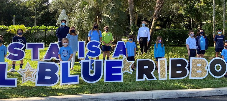 Students, principal Lisa Figueredo (middle left) and pastor, Father Alejandro Rodriguez Artola (rear right)pose amid the Blue Ribbon sign on the lawn of St. Thomas the Apostle School in Miami, which was one of two archdiocesan schools honored by the U.S. Department of Education in 2020.