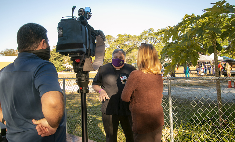 El P. José Espino, párroco de la iglesia San Lázaro en Hialeah, habla con una reportera de Telemundo 51 mientras la vacunación está en marcha detrás de él, el 20 de enero de 2021.