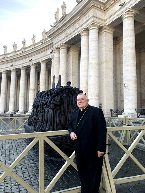 El Arzobispo Thomas Wenski posa frente a la escultura Angels Unawares que se exhibe en la Plaza de San Pedro en Roma, en febrero de 2020, durante su visita ad limina con el Papa Francisco. Una réplica estará expuesta en el Bayfront Park de Miami, del 9 de febrero al 8 de abril de 2021.