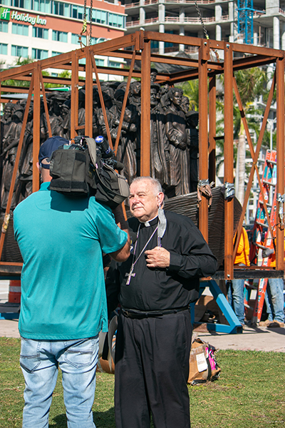 Archbishop Thomas Wenski gives interviews to local media as workers from Florida Lemark unload and install the sculpture "Angels Unawares" in Miami's Bayfront Park, where it will be on display through April 8, 2021. The installation took place Feb. 9, 2021.