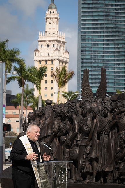 Con la Torre de la Libertad en el fondo y la escultura delante de él, el Arzobispo Thomas Wenski habla en la ceremonia de bendición de “Ángeles sin saberlo”, en Bayfront Park, el 10 de febrero de 2021. El Arzobispo calificó a Miami como la “Ellis Island del Sur.” La escultura, un tributo a los migrantes y refugiados, permanecerá en el parque hasta el 8 de abril de 2021.