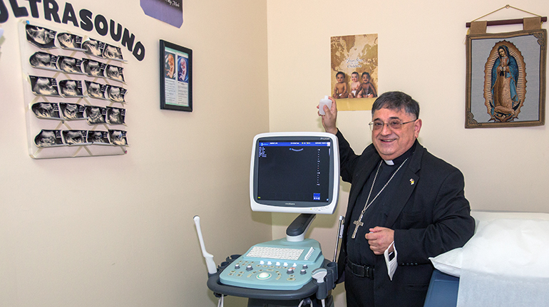 Auxiliary Bishop Enrique Delgado sprinkles holy water on the new ultrasound machine at the North Broward Pregnancy Help Center, March 3, 2021. The more advanced machine was donated by Mario Labella, CEO of Strena Medical Products in Doral, at the request of St. Katharine Drexel's Knights of Columbus council.