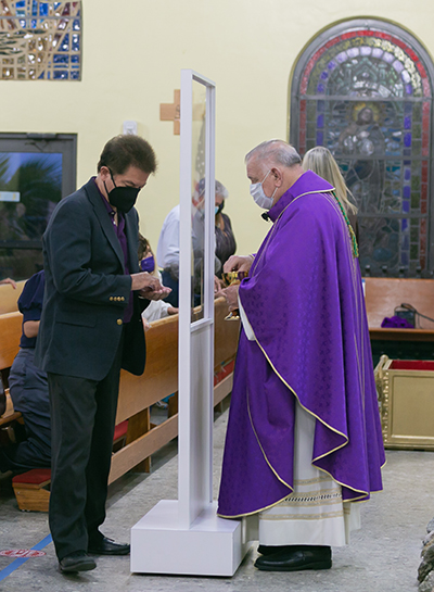 Archbishop Thomas Wenski distributes Communion Peter Routsis-Arroyo, CEO of Catholic Charities, during the agency's 90th anniversary Mass at St. Joachim Church in Miami, March 26, 2021. A temporary barrier helps maintain social distance during the COVID-19 pandemic.